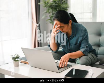 Stressed Asian woman using laptop at home and touching forehead from headache, concept of remote work pressure, burnout, digital fatigue, online work, Stock Photo