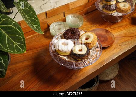 Selection of gourmet donuts with chocolate sprinkles, powdered sugar, and cream topping displayed on a glass cake stand over a warm wooden table in na Stock Photo