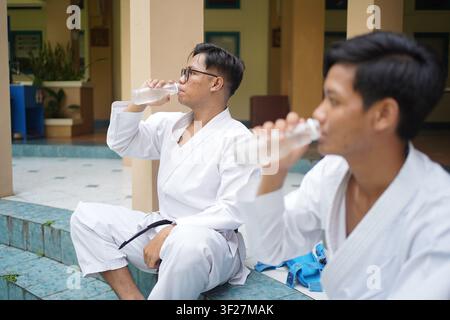 Two Asian Indonesian karate athletes drinking water while resting after martial arts training session, highlighting hydration and recovery. Stock Photo