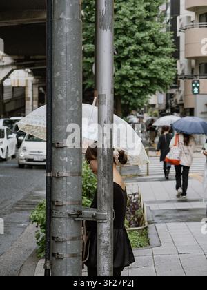 Woman with transparent umbrella behind utility poles on a rainy street in Shibuya, Tokyo, with pedestrians crossing in the background Stock Photo