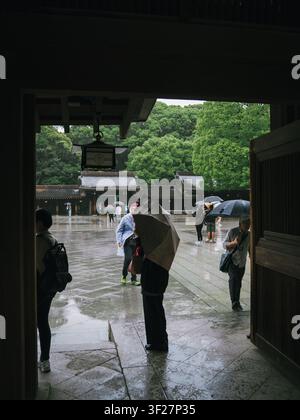 Visitors with umbrellas in the rain-soaked courtyard of Meiji Shrine seen through a wooden gate, Tokyo, Japan Stock Photo