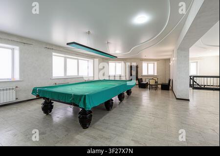 well-lit game room features a pool table covered in green cloth. The room includes seating, windows, and unique ceiling designs Stock Photo