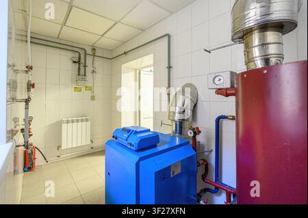 utility room featuring a blue boiler, red tank, pipes, white tiled walls, and a tiled floor. A door opening is visible on the back wall Stock Photo