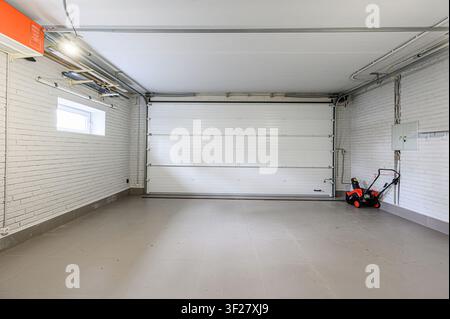 clean, well-lit garage interior with a white sectional door, brick walls, tiled floor, and a red snow blower Stock Photo