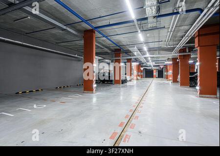 well-lit indoor parking garage with orange pillars, concrete flooring and ceiling, visible pipes, and marked parking spaces. Several cars are parked i Stock Photo