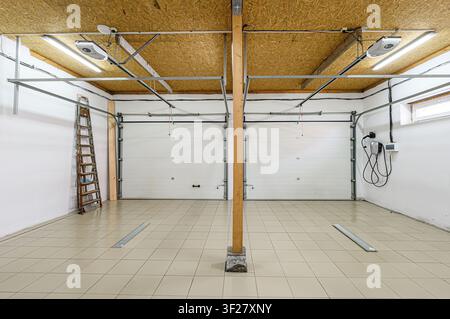 well-lit garage interior with two closed white doors, a wooden supporting beam, tiled floor, and a ladder on the side Stock Photo