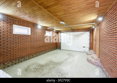 interior garage space featuring brick walls, a light wooden plank ceiling, concrete floor, a white garage door, and two small windows Stock Photo