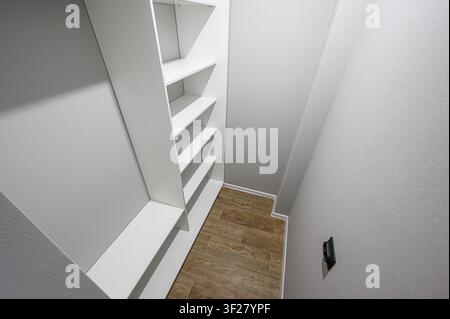 narrow, empty walk-in closet features white walls and shelves. The floor is tiled in a wood-like pattern. A wall switch is visible on the right Stock Photo