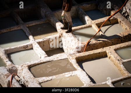 High angle daytime view of a tannery worker standing on narrow edges between pale tanning vats, with rubber boots, gloves and an orange hose at Chouar Stock Photo