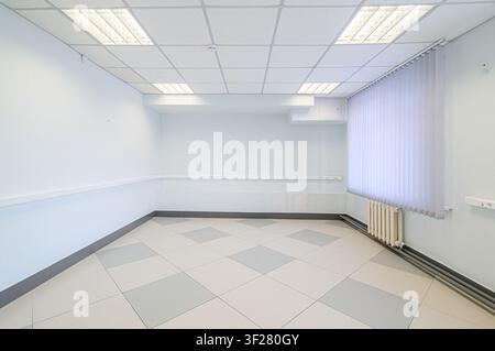 empty, brightly lit room with white walls, tiled floors, and vertical blinds on the window. The room has a drop ceiling and a radiator Stock Photo
