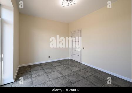 Empty room with beige walls, gray tiled floor, a white door with horizontal lines, and a modern ceiling light fixture. Natural light enters from a win Stock Photo