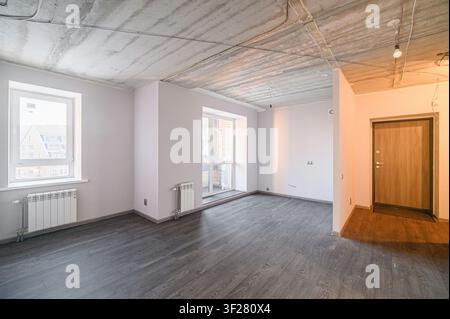 An empty room under renovation with grey flooring, white walls, and a concrete ceiling with visible wiring. Windows with radiators are present. A wood Stock Photo