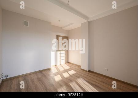 An empty room with light beige walls, wooden floor, and a light-colored door. Sunlight streams in, casting shadows across the floor Stock Photo