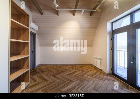 Bright attic room with herringbone wood floor, light wood paneling on the ceiling, a bookcase, and doors to a balcony. Clean, modern design Stock Photo