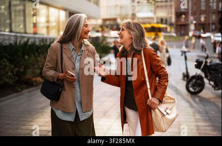 Animated Street Conversation Between Two Stylish Silver Haired Women Walking Together in a Busy Urban District Carrying Handbags in Warm Afternoon Stock Photo
