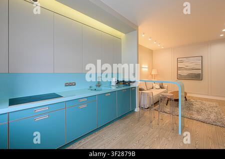 Modern kitchen with blue cabinets and countertops, seamlessly connected to a living room with sofa and stools. Warm lighting enhances the space Stock Photo
