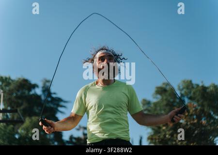 man actively jumping rope in an outdoor setting on a sunny day, surrounded by trees, wearing a green t-shirt and black shorts, captured from a low angle that emphasizes his energetic motion. Stock Photo