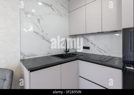 Modern kitchen corner with white cabinets, dark countertops, marble backsplash, and a black sink and faucet. Clean and sleek design Stock Photo