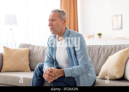 Worried Elderly Caucasian Man Sitting On Couch At Home, Focused On Thoughts, Healthcare Concept Stock Photo