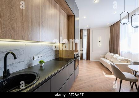 Modern apartment featuring a kitchen with wood cabinets, black countertops, marble backsplash, and open living area with sofa and dining table Stock Photo