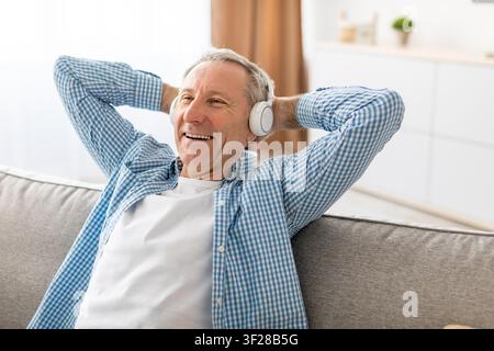 Happy Elderly Caucasian Man Relaxing On Sofa Listening To Music With Headphones, Home Living Room Stock Photo