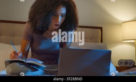 African American woman student girl writing notes looking in laptop on bed at night dark bedroom ethnic female look in computer write watch video Stock Photo