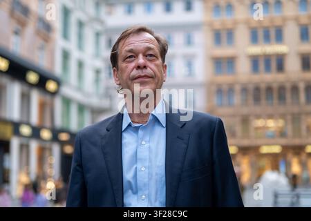 A middle-aged man wearing a dark blazer and light blue shirt stands outdoors in an urban street setting. The background shows softly blurred buildings Stock Photo