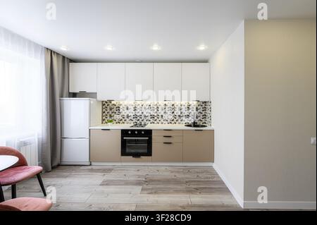 modern kitchen with white ribbed upper cabinets, beige lower cabinets, and geometric tile backsplash is shown. Pink chairs are at the edge Stock Photo