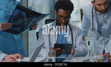 Professional multiethnic medical team of experienced doctors discussing x-ray using digital tablet laptop talking about diagnosis healthcare briefing Stock Photo