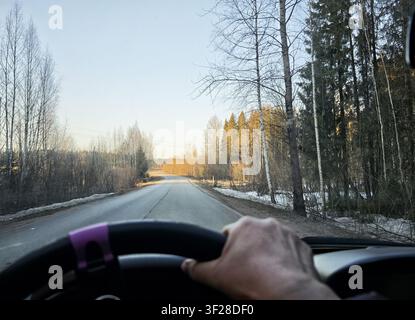 A person drives on a quiet road in early spring, with trees lining the sides and sunlight shining in the distance. Stock Photo