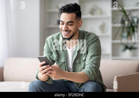 Smiling Young Asian Man Using Smartphone At Home, Relaxed Indoor Atmosphere Stock Photo