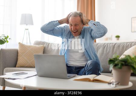 Happy Elderly Caucasian Man Celebrating With Hands On Head While Using Laptop At Home Stock Photo