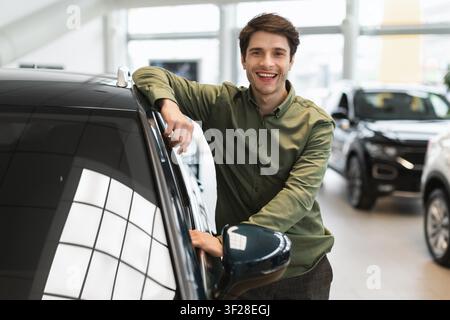 Smiling Young Caucasian Man Standing Next To Car In Showroom, Car Sales Concept Stock Photo