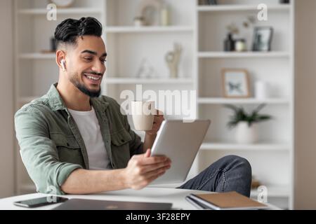 Smiling Hispanic Male Using Tablet While Holding Coffee Mug At Home, Relaxing Indoor Stock Photo
