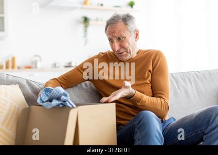 Worried Elderly Caucasian Man Sitting On Sofa, Opening Package At Home, Surprise Expression Stock Photo