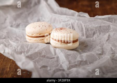 beige macaroons with chocolate filling, coffee or cappuccino flavor and cocoa powder on paper, wooden background. Close up shot Stock Photo