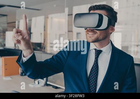 Young financier experiencing immersive virtual reality technology wearing VR glasses, interacting with 360 degrees headset user interface, while standing in his office, selects content in 3D menu Stock Photo