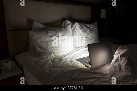 Open laptop glowing on unmade bed at night, dark bedroom interior, remote work overtime insomnia burnout concept, technology dependency, freelance lifestyle mental fatigue copyspace Stock Photo