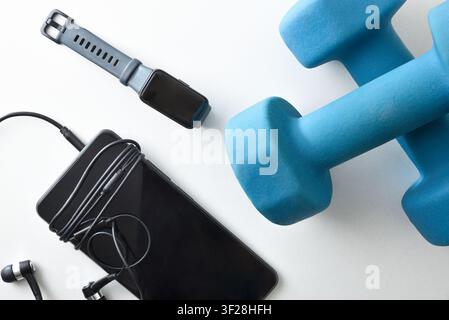 Sports and leisure items on a white tabletop, such as blue neoprene dumbbells, a smartwatch, and a phone with headphones. Top view. Stock Photo
