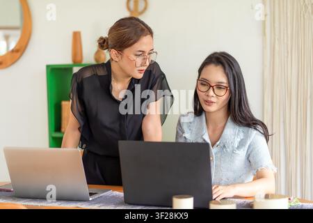 Two Asian professional women collaborating on a laptop project in a bright modern workspace. Stock Photo