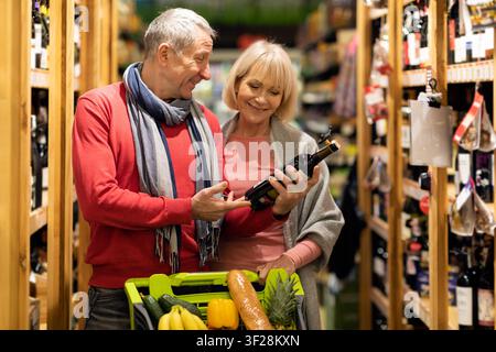 Happy Elderly Caucasian Couple Shopping Together In Supermarket, Choosing Wine Bottle Stock Photo
