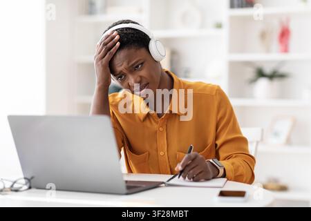 Worried Black Woman Wearing Headphones Taking Notes While Working At Laptop, Home Office Stock Photo
