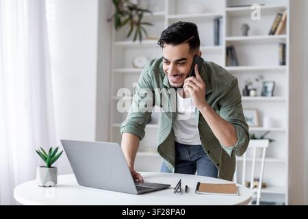 Smiling Young Hispanic Man Using Laptop While Talking On Phone At Home Office Desk Stock Photo