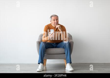 Thoughtful Elderly Caucasian Man Using Smartphone While Sitting In Cozy Armchair, Senior Lifestyle Concept Stock Photo