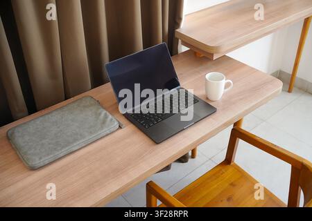 Clean home workspace featuring a laptop, coffee mug, and laptop sleeve on a wooden desk with curtains, representing minimalism, productivity, and a co Stock Photo