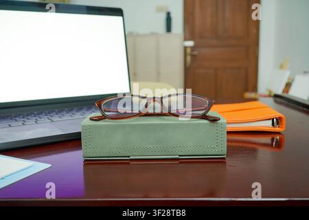 Stylish eyeglasses placed on a glasses case beside laptop and orange notebook on wooden desk, creating a productive home office workspace. Stock Photo