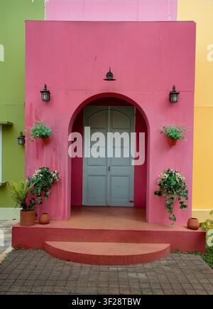 A vibrant pink arched doorway framed by hanging and potted plants, black lanterns and pastel green and yellow walls. Charming colorful facade evokes w Stock Photo