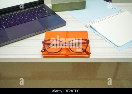 Stylish eyeglasses placed on orange notebook beside laptop and open notepad on white desk, ideal for study and remote work concepts. Stock Photo