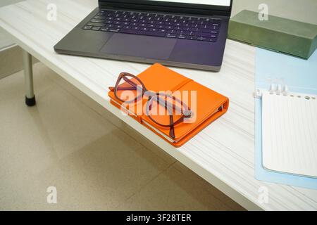 Stylish reading glasses placed on orange notebook beside laptop and open notepad on white desk for study, planning, and remote work concepts. Stock Photo