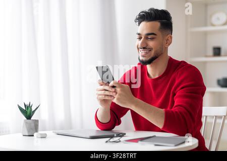 Smiling Young Middle Eastern Man Using Smartphone At Home Office, Business Concept Stock Photo